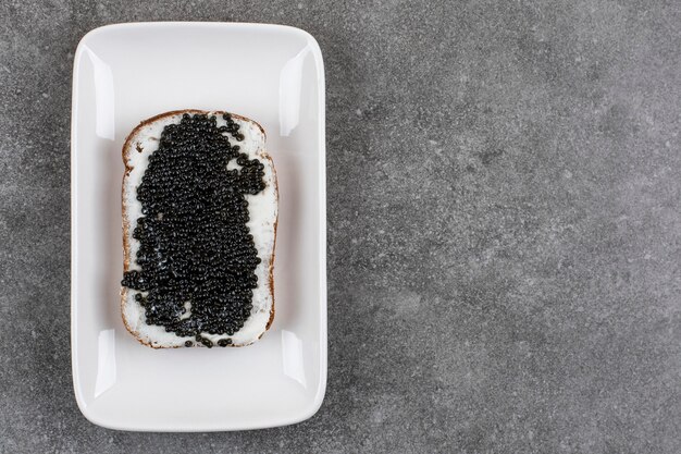 Вкусный бутерброд с черной икрой на белой тарелке над серой поверхностью