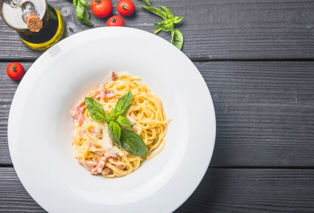 Вкусные спагетти в белой тарелке с оливковым маслом; помидоры и листья базилика на деревянном столе