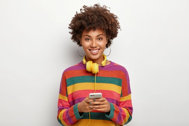 Delighted African American teenage girl holds mobile phone connected to headphones