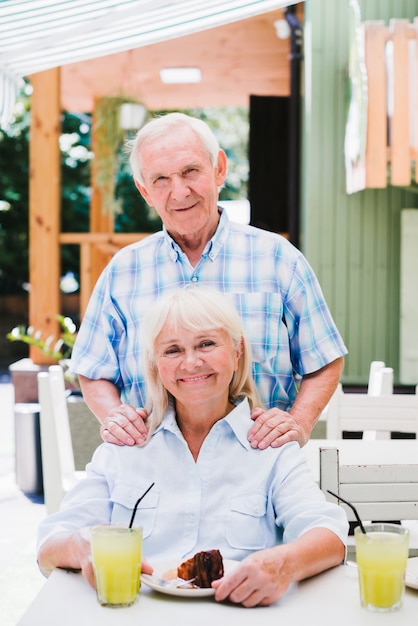 Delighted aged couple hugging in cafe on terrace enjoying refreshing drink and cake