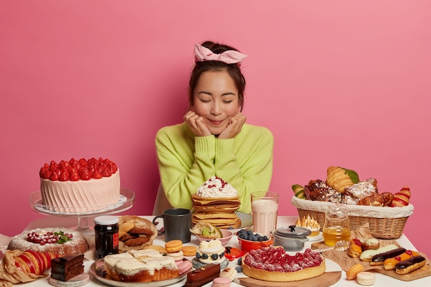 Free photo delighted asian woman wears headband and green jumper, holds chin, has good appetite, eats sweet food, fruit cakes, comes on birthday party, isolated on pink wall