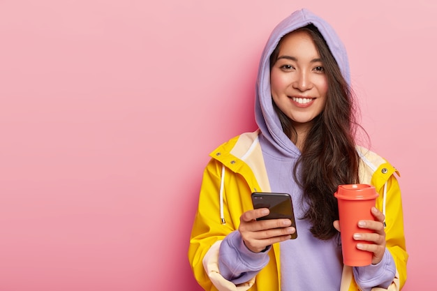 Delighted attractive Asian woman wears casual clothes, raincoat, sends message via cell phone, drinks aromatic beverage from thermos