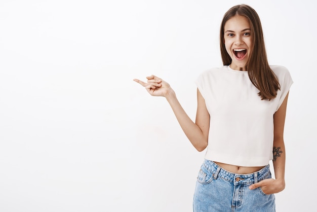 Delighted attractive caucasian female brunette with tattoo on arm in white t-shirt smiling joyfully with opened mouth from fascination and excitement holding hand in pocket pointing left at copy space