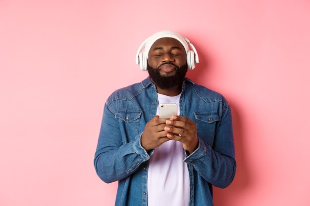 Delighted Black man enjoying awesome music, listening songs in headphones and holding smartphone, looking ecstatic, standing over pink background