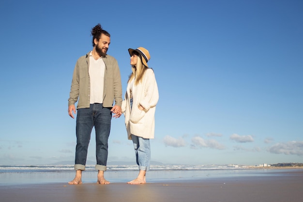 Delighted couple walking on beach. Bearded man and woman in casual clothes looking at each other, holding hands. Love, vacation, affection concept