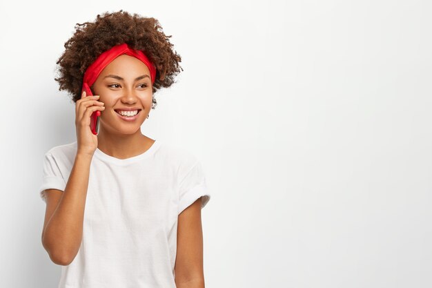Обрадованная кудрявая афро-женщина держит мобильный телефон возле уха, наслаждается приятной беседой, позитивно улыбается