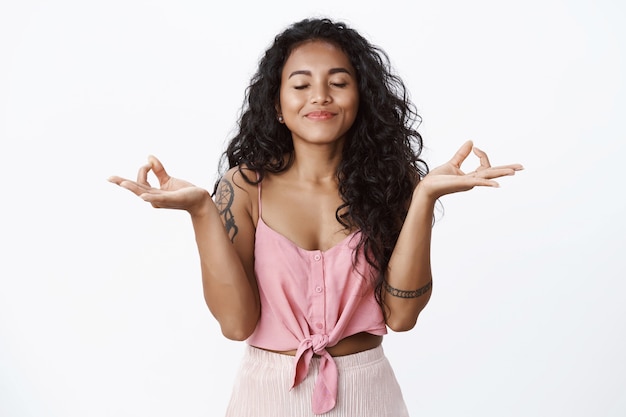 Delighted curly-haired girl with tattoos meditating, raise hands in zen gesture, practice yoga, close eyes and smiling happy, inhale fresh air, calm down relieved, white wall
