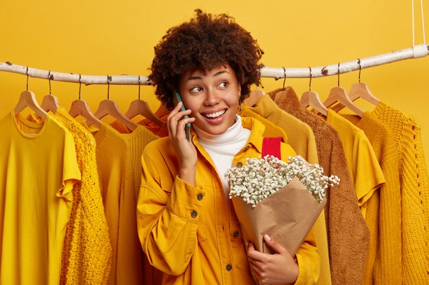 delighted curly woman turns away, calls via cellular, shares impressions after shopping day, poses with bouquet of flowers