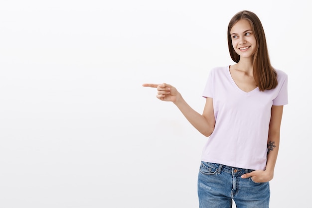 delighted cute caucasian female brunette with awesome tattoo on arm pointing and looking left with appreciation and thankful expression smiling joyfully posing against white wall