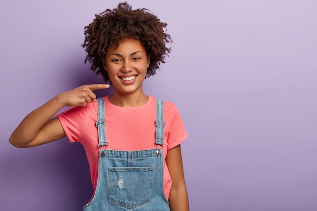 Felice ragazza dalla pelle scura sorride ampiamente, mostra denti bianchi perfetti, indossa una maglietta casual rosa e una tuta di jeans, è di buon umore, isolato sul muro viola