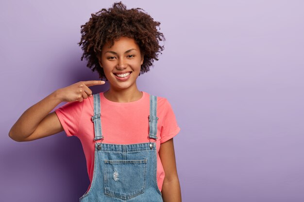 Delighted dark skinned girl smiles broadly, shows white perfect teeth, wears pink casual t shirt and denim overalls, being in good mood, isolated on purple studio wall