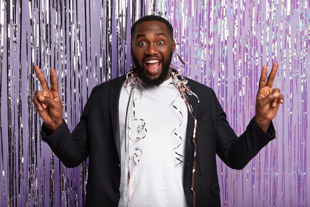 Delighted dark skinned man raises hands, shows peace gesture, laughs happily, has serpentine on body, dressed in elegant suit, expresses positive emotions, stands over decorated purple wall