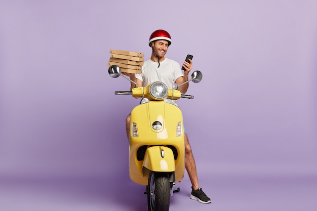 Delighted deliveryman driving scooter while holding pizza boxes