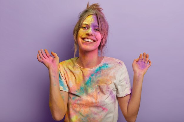 Delighted European woman with carefree expression, raises hands, smeared with colorful dyes, wears white t shirt, smiles gladfully, celebrates Holi fest, paint party isolated over purple wall.