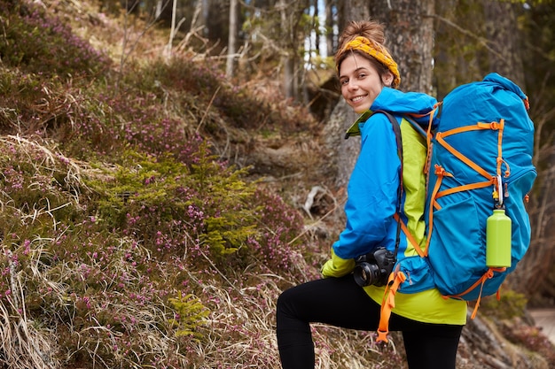 Delighted female camper carries backpack, professional camera