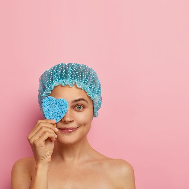 Delighted female model covers eye with heart shaped sponge, wears showercap, stands naked against pink wall, wipes makeup, has hygienic procedures, cares about face skin, models indoor