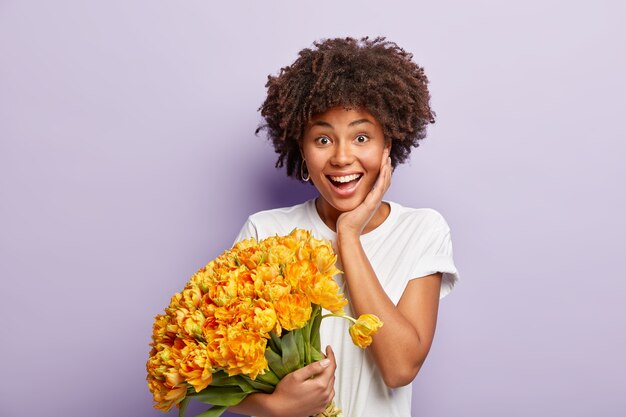 이빨 미소, 선명한 머리카락, 남자 친구로부터 행복한 제안을받은 기쁘게 여성은 보라색 벽에 고립 된 노란색 꽃의 멋진 꽃다발을 보유하고 있습니다. 긍정적 인 감정과 감정 개념