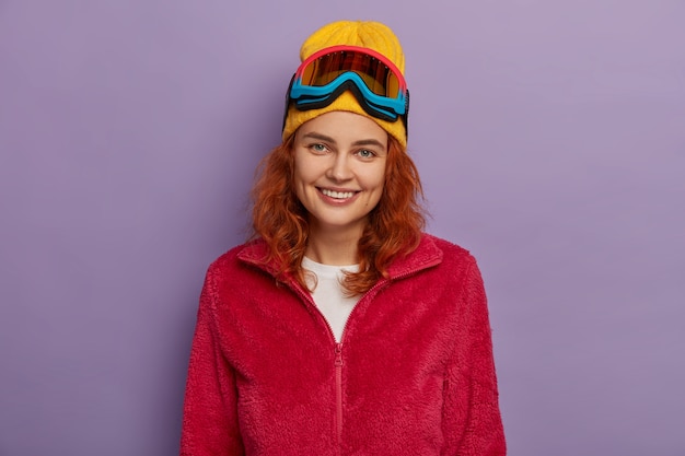 Delighted ginger woman with appealing appearance, wears yellow hat and red soft jacket, has pleasant smile on face, looks directly at camera, isolated over purple background.