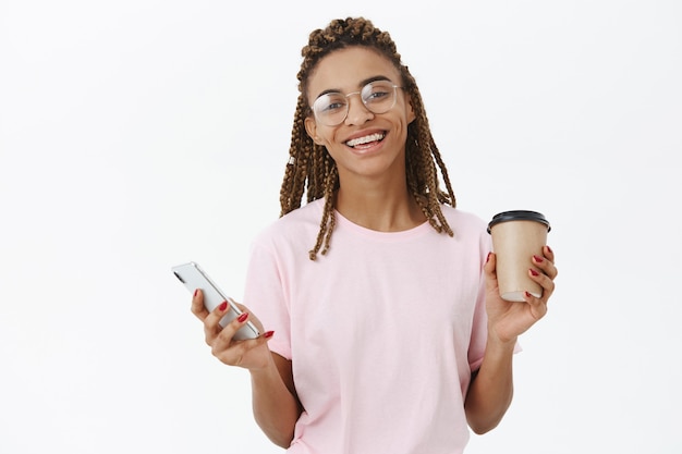 Delighted happy and amused charming african american girl with dreads in glasses holding paper cup of coffee and smartphone