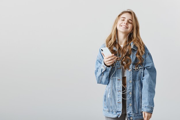 Delighted happy blond girl enjoying awesome new song in earphones, listening music, holding smartphone