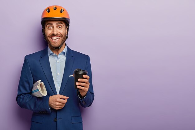 Delighted happy male construction worker has coffee break after work, wears hardhat and elegant suit, smiles positively