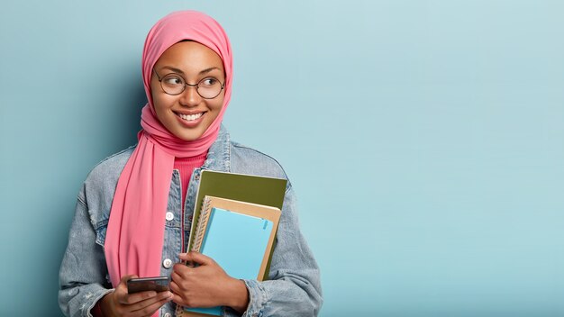 Delighted happy Muslim student types messages on cell phone, carries notepad, focused aside with joyful expression, wears jean jacket, isolated against blue wall, reads interesting article