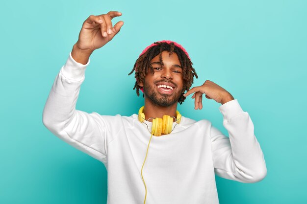 Delighted happy teenager with dreadlocks, raises arms, feels joy as listens favourite music via headphones, moves in rhythms of song, wears white jumper