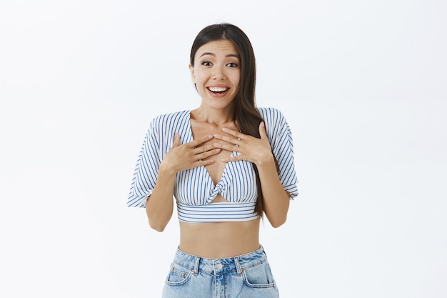 delighted happy and touched feminine dark-haired asian woman in cropped blouse holding palms on chest