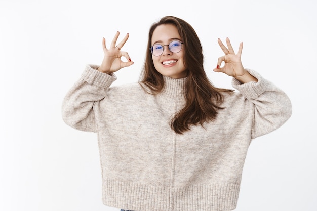 Delighted happy young female in glasses and stylish sweater raising hands, showing okay, smiling pleased giving opinion about awesome trip, sharing positive emotions and impressions.