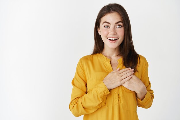 Delighted and happy young woman looking with admiration and joy, holding hands on heart in appreciation of gift, standing touched over white wall.