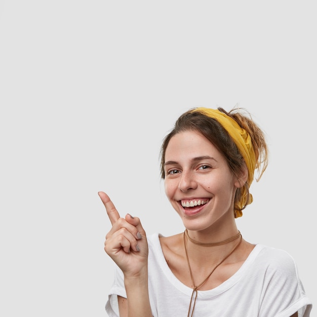 Delighted joyful woman points at upper left corner, enjoys spending free time at shopping, smiles broadly