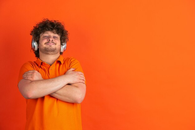 С удовольствием слушаю музыку. Монохромный портрет кавказского молодого человека на оранжевой стене. Красивая мужская фигурная модель в стиле casual. Понятие о человеческих эмоциях, выражении лица.