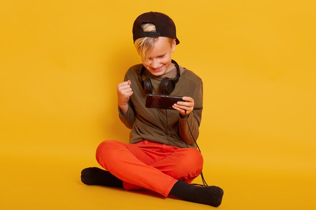 delighted little boy dresses orange trousers and green shirt playing video game on cellphone and clenching fist while sitting on floor with crossed legs isolated on yellow