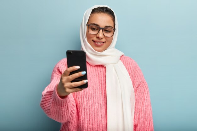Восхищенная мусульманка с нежной улыбкой, держит перед собой мобильный телефон, делает селфи-портрет, носит оптические прямоугольные очки и вуаль, имеет маникюр. Время делать фото