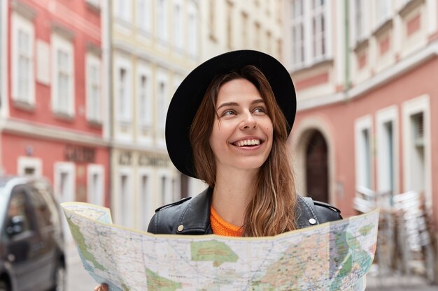 Delighted optimistic female toursit finds place on map, walks in city center during summer trip, wears stylish black hat, poses against urban setting