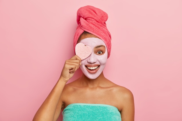 Delighted pleased female model holds cosmetic sponge, applies facial clay mask which suits her skin, has cosmetic procedures in bathroom