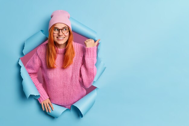 Delighted pleased ginger woman points away with thumb pointing at copy space