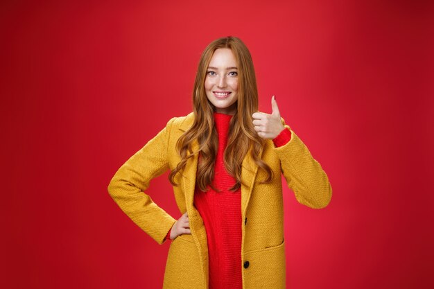 Обрадованная и довольная женщина-рыжая покупательница показывает большие пальцы в знак одобрения и одобрения, довольная отличным качеством нового желтого пальто, стоящего на красном фоне, широко улыбаясь.