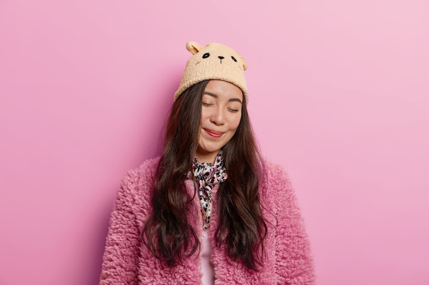 Free photo delighted tender woman stands with closed eyes and charming smile on face, wears hat with ears and rosy fur coat, turns face from camera
