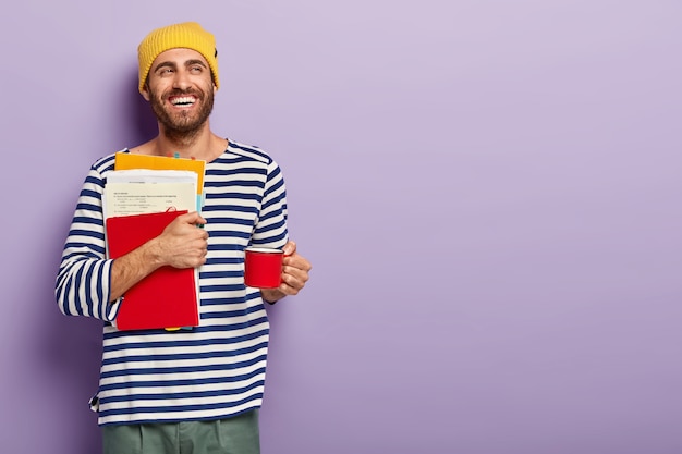 Foto gratuita felice con la barba lunga giovane studente tiene documenti e blocco note rosso, tiene la tazza con una bevanda calda, ha una pausa caffè, essere di buon umore, guarda da parte con un ampio sorriso, isolato su sfondo viola