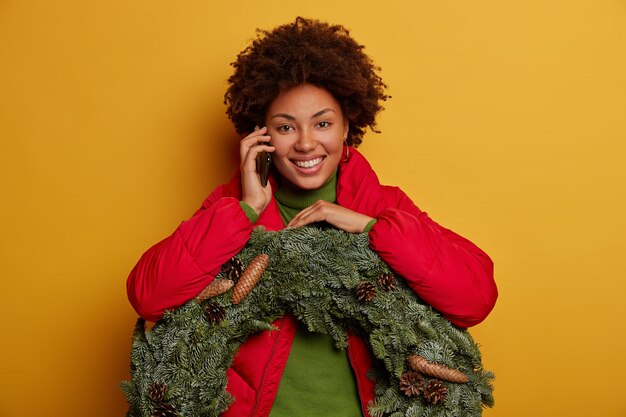 Delighted woman calls via cellphone, has festive mood on Christmas Eve, tells latest news, carries spruce wreath with pine cones, smiles broadly isolated on yellow wall.