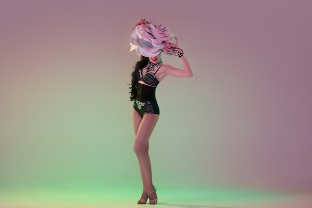 Delighted. Young female dancer with huge floral hats in neon light on gradient wall.