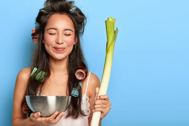 Delighted young female model with hair curlers, carries green leek and bowl, makes salad of healthy ingredients, keeps eyes shut