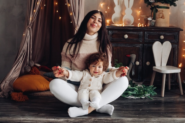 Delighted young lady sitting cross-legged on floor while holding a cute kid on her lap and smiling