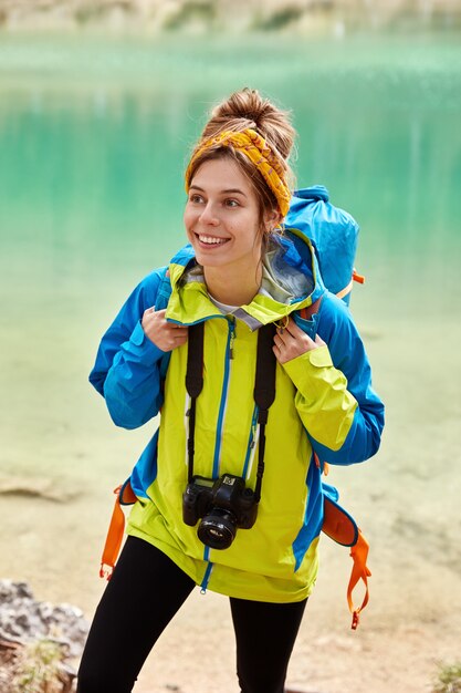 Delighted young woman tourist has combed hair, wears scarf on head, colorful anorak, holds camera