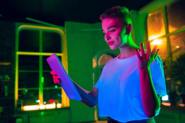 Delightful. Cinematic portrait of stylish woman in neon lighted interior. Toned like cinema effects, bright neoned colors. Caucasian model using tablet in colorful lights indoors. Youth culture.