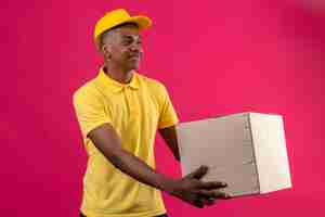 Free photo delivery african american man in yellow polo shirt and cap giving a paper package to a customer smiling friendly on isolated pink