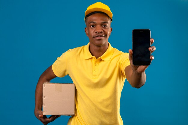 Афро-американский мужчина доставки в желтой рубашке поло и кепке, держащий коробку, показывает мобильный телефон с улыбкой на лице, стоящий на синем