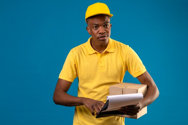 Афро-американский мужчина доставки в желтой рубашке поло и кепке держит картонную коробку и буфер обмена, сосредоточенный на задаче, стоящей на изолированном синем