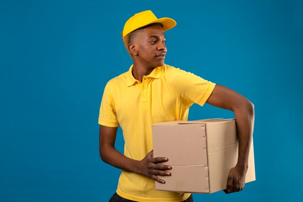 Delivery african american man in yellow polo shirt and cap holding cardboard boxes looking aside with smile on face standing on blue